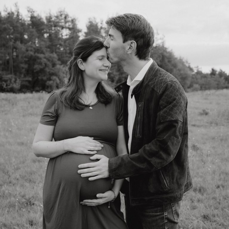 Schwangerschaft Fotoshootings Schwangere Frau in einem Kleid steht mit einem Mann in einer Feldlandschaft.